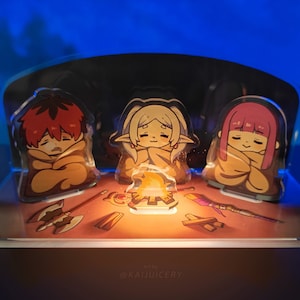 May include: Three anime characters are bundled up in blankets around a glowing campfire. The characters are made of clear acrylic and are part of a larger scene with a brown background. The text "@KAJUICERY" is at the bottom of the image.