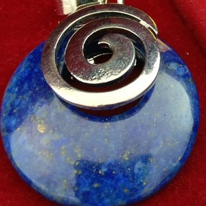 Puede incluir: Un colgante de lapislázuli azul con un diseño de espiral plateado. El colgante está en una caja de terciopelo rojo.