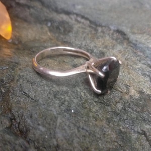 May include: A silver ring with a large, black gemstone. The ring has a simple band and a four-prong setting that holds the dark, faceted stone. The ring is set against a textured, green-gray surface.