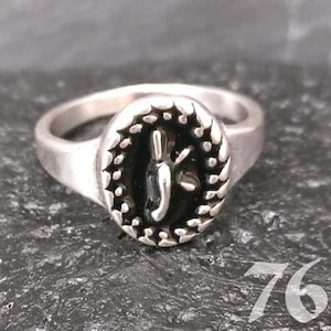 May include: A silver ring with an oval, black center featuring a stylized design. The ring has a textured border and a smooth band. The number "76" is visible in the lower right corner. The ring is displayed on a dark, textured surface.