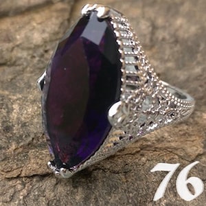 May include: A silver-toned ring with a large, marquise-cut, deep purple gemstone. The ring has an ornate, filigree design on the band and around the stone. The number "76" is visible in the lower right corner.