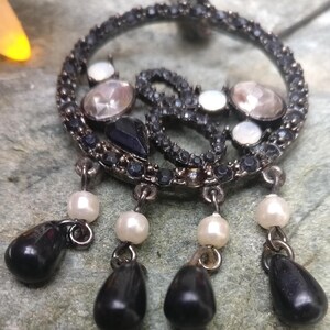 May include: A silver-toned pendant with a circular design, adorned with clear and black gemstones, and white pearls. The pendant has three black teardrop-shaped dangles. The pendant is on a textured, gray surface.
