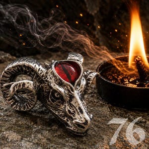 May include: A silver-toned metal ram head figurine featuring a prominent, oval-shaped red eye with a vertical black pupil. The ram head is positioned next to a small, round black container with a burning flame and visible smoke trails.