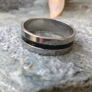 May include: A silver ring with a black inlay. The ring is made of a shiny metal and has a flat band design. The black inlay is a thin stripe that runs around the center of the ring. The ring is sitting on a gray stone surface.