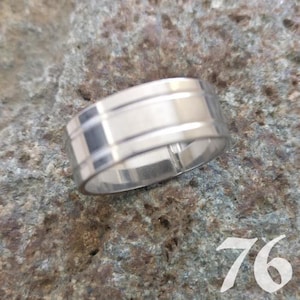 Könnte beinhalten: Ein silberfarbener Metallring mit glatter, polierter Oberfläche und zwei dünnen, horizontal eingravierten Linien, die das Band umkreisen. Der Ring hat ein flaches Profil und liegt auf einer strukturierten, grauen Steinoberfläche mit der Zahl 76.