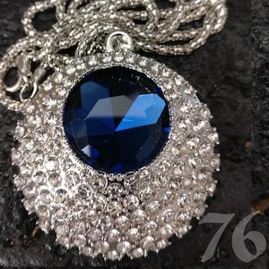 Puede incluir: Un collar plateado con un colgante grande y redondo. El colgante presenta una piedra preciosa azul oscuro facetada, rodeada de pequeños cristales transparentes. La cadena es de diseño delicado y enlazado.