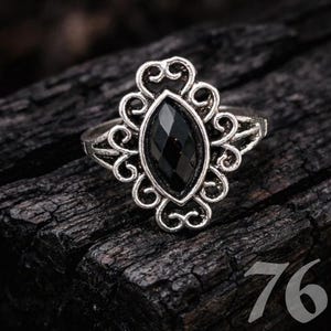 May include: Silver ring with a large, marquise-cut black gemstone. The ring has an ornate, filigree design around the stone and on the band. The number "76" is visible in the lower right corner.