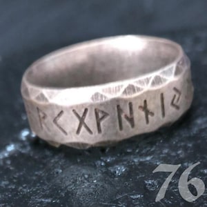 May include: A silver-colored ring with etched runic symbols. The ring has a textured surface with a geometric pattern along the top edge. The runes are carved into the band, adding a unique, ancient aesthetic.