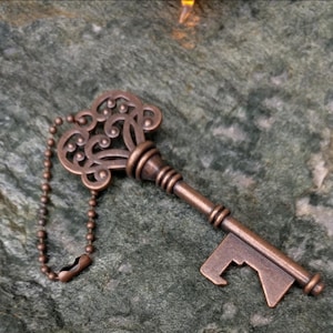 May include: A bronze-coloured key-shaped bottle opener with an ornate, filigree design on the key head. The opener has a small chain attached and rests on a textured, green-grey surface. The key's design is vintage-inspired.