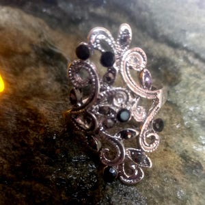 Puede incluir: Anillo plateado con un diseño ornamental y arremolinado. El anillo está adornado con piedras preciosas negras y moradas. El intrincado trabajo en metal presenta patrones florales y de volutas, creando una estética vintage o antigua. El anillo se encuentra sobre un fondo oscuro y texturizado.