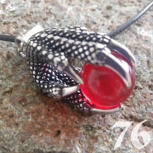 May include: A silver-colored claw pendant grasping a red orb. The claw has a textured, dotted pattern and sharp, polished talons. The pendant is suspended from a black cord.