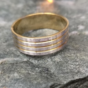 May include: A gold and silver-toned ring with a brushed metal finish. The ring features four horizontal silver bands separated by gold bands. The ring is set against a textured, gray surface.