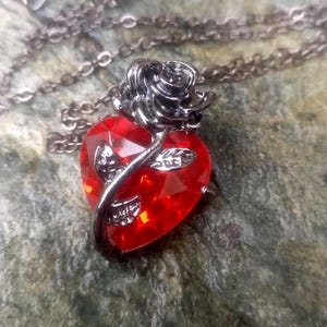 May include: A silver necklace with a red heart-shaped pendant. A silver rose and stem design adorns the heart. The chain is silver. This pendant is a romantic and striking piece of jewellery.