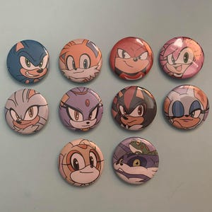 May include: Ten circular badges showcasing characters from the Sonic the Hedgehog series. Each badge features a different character's face, with bright colours and detailed artwork. The badges are arranged on a light blue surface.