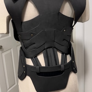 May include: Black leather corset with a structured design and a back panel with cutouts. The corset has a series of straps and buckles around the waist.