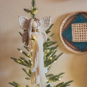 May include: A handmade macrame angel Christmas tree topper with a wooden head, gold accents, and long, flowing cream-colored yarn. The angel has large, detailed wings and is placed on top of a green Christmas tree with fairy lights.