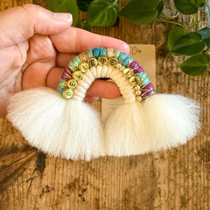 May include: A handmade rainbow-shaped wall hanging with the words "BELIEVE IT" spelled out in gold letters. The rainbow features a cream-colored arch, with blue, purple, and gold accents. Fluffy white tassels form the base of the rainbow.