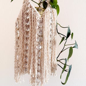 Puede incluir: Colgador de plantas de macramé color crema con detalles de cuentas de cristal transparente. El soporte para plantas tiene un fondo con flecos y está suspendido de una cuerda trenzada. Las vides verdes y frondosas caen en cascada desde la parte superior, creando una estética bohemia.