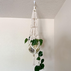 Puede incluir: Un colgador de plantas de macramé con una maceta de bola de discoteca que contiene una planta verde. El macramé blanquecino tiene un diseño en capas con detalles dorados y una borla. Las hojas verdes de la planta caen, creando una exhibición interior decorativa.