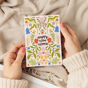 May include: A rectangular greeting card with a light pink background featuring a colorful floral illustration and a small house icon with a heart. The text Happy New Home is printed in the center in black script. The design includes blue, red, yellow, and pink flowers with green leaves and stems.