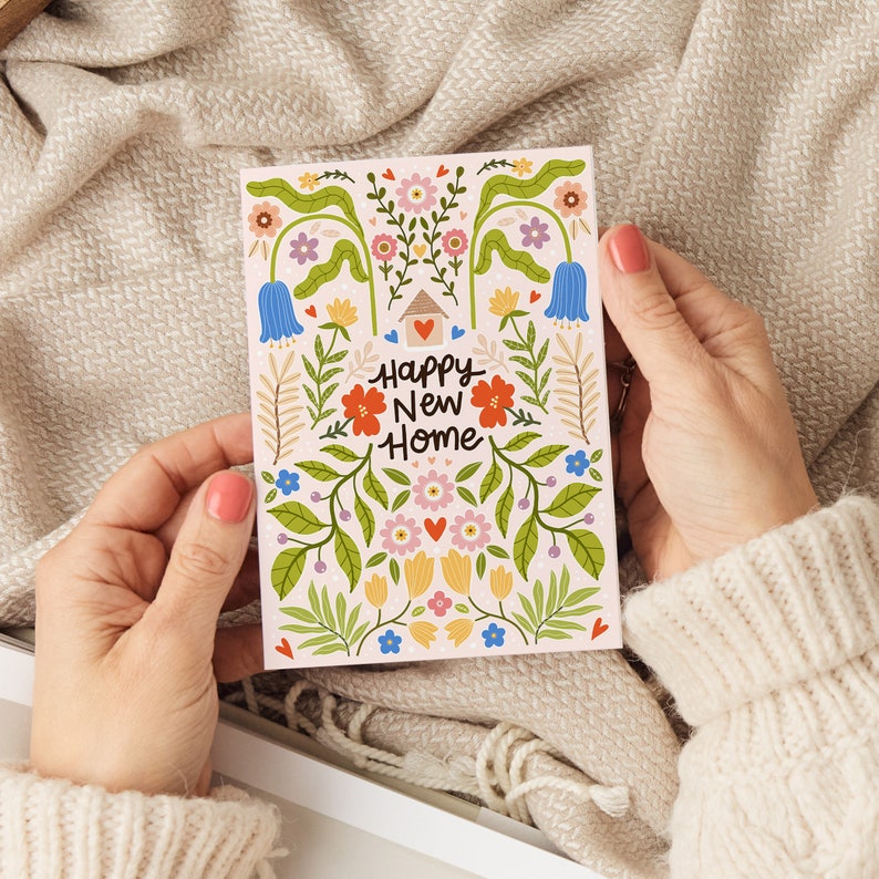 May include: A rectangular greeting card with a light pink background featuring a colorful floral illustration and a small house icon with a heart. The text Happy New Home is printed in the center in black script. The design includes blue, red, yellow, and pink flowers with green leaves and stems.