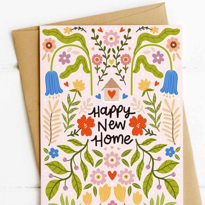 May include: A greeting card with a light pink background featuring a colorful floral pattern, green leaves, small hearts, and a central house icon with a red heart. The text Happy New Home is printed in dark brown in the center of the card.