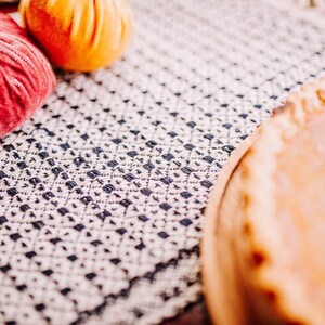 Op de afbeelding: Een close-up van een pompoentaart op een wit en blauw gepatroneerd tafelkleed. De taart zit in een houten taartvorm en heeft een goudbruine korst. Er zijn twee decoratieve pompoenen op de voorgrond, een rode en een oranje.
