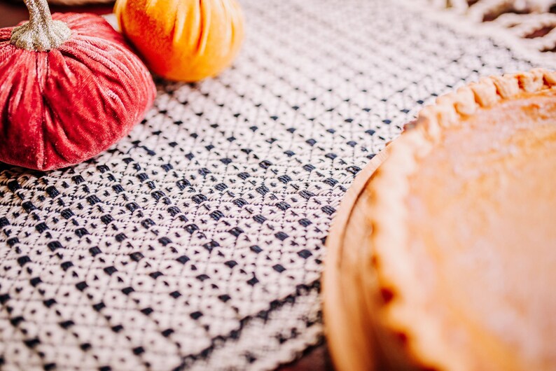 Op de afbeelding: Een close-up van een pompoentaart op een wit en blauw gepatroneerd tafelkleed. De taart zit in een houten taartvorm en heeft een goudbruine korst. Er zijn twee decoratieve pompoenen op de voorgrond, een rode en een oranje.