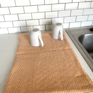 May include: A mustard yellow and white woven dish drying mat with two white ceramic mugs. The mat is rectangular and sits on a countertop next to a stainless steel sink. The background features white subway tile.