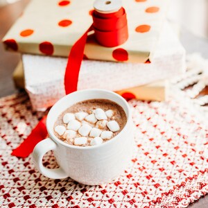 Op de afbeelding: Een witte mok gevuld met warme chocolademelk, bedekt met mini marshmallows. De mok staat op een rood-wit geblokt placemat. Op de achtergrond staat een stapel ingepakte cadeaus met rood lint.