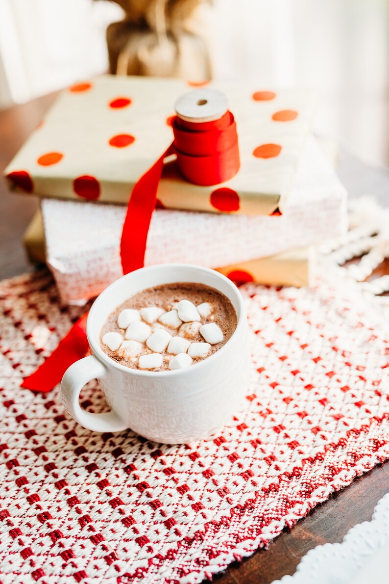 Op de afbeelding: Een witte mok gevuld met warme chocolademelk, bedekt met mini marshmallows. De mok staat op een rood-wit geblokt placemat. Op de achtergrond staat een stapel ingepakte cadeaus met rood lint.