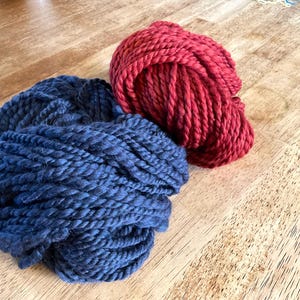 May include: Two balls of yarn, one deep blue and one red, sit on a wooden surface. The yarn appears thick and textured, suggesting a chunky knit or crochet project. The colors are rich and saturated.