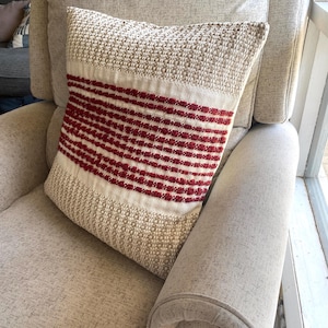 May include: A decorative pillow with a woven texture and a rectangular shape. The pillow features a cream-colored top and bottom with a central band of horizontal red stripes. It is placed on a beige upholstered armchair.