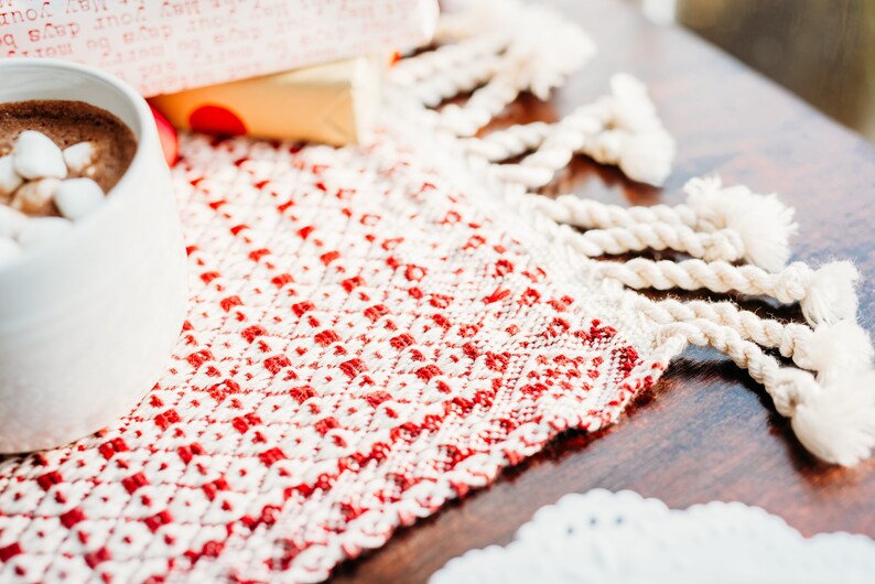 Op de afbeelding: Een witte mok gevuld met warme chocolademelk met marshmallows erop, staat op een rood-wit geweven placemat met franjes.