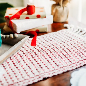 Op de afbeelding: Een rode en witte geweven tafelrunner met franjes aan de randen. De runner ligt op een houten tafel met kleine kunstkerstbomen in een houten dienblad.