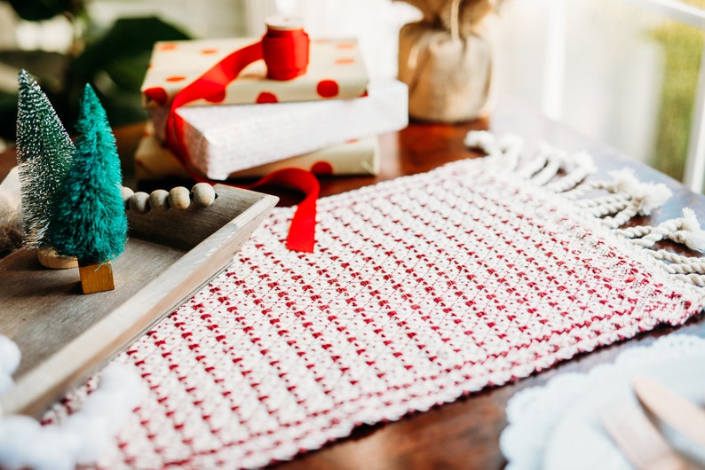 Op de afbeelding: Een rode en witte geweven tafelrunner met franjes aan de randen. De runner ligt op een houten tafel met kleine kunstkerstbomen in een houten dienblad.