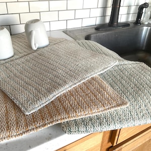 May include: Three rectangular dish drying mats with a thick, braided texture. One mat is a light blue and white blend, another is a warm tan and white mix, and the third is a soft sage green and white combination. Two white ceramic mugs sit on top.