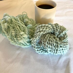 May include: Two skeins of chunky, hand-dyed yarn in shades of pale green and blue-green, with a textured, knotted appearance. A beige mug filled with dark coffee sits nearby, suggesting a cozy crafting scene.