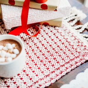 Op de afbeelding: Een witte mok gevuld met warme chocolademelk met marshmallows erop staat op een rood-wit gepatroneerd placemat. De mok staat naast een stapel ingepakte cadeaus die vastgebonden zijn met een rood lint.