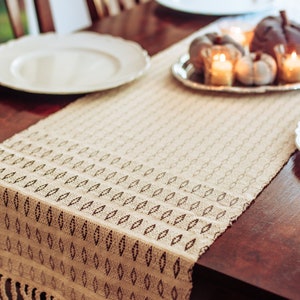 May include: A beige crocheted table runner with a geometric pattern and fringe edges. The runner is on a wooden table with two white plates and a silver tray with small pumpkins and candles.