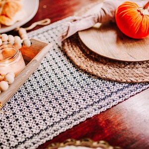 Op de afbeelding: Een houten dienblad met een wit en zwart gepatroneerde loper, een brandende kaars en herfstdecoraties, waaronder pompoenen, bessen en een fluwelen pompoen.