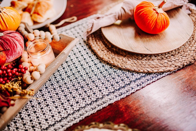 Op de afbeelding: Een houten dienblad met een wit en zwart gepatroneerde loper, een brandende kaars en herfstdecoraties, waaronder pompoenen, bessen en een fluwelen pompoen.