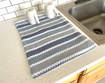 May include: A rectangular dish drying mat with alternating stripes of white, light blue, and dark blue. Two white mugs sit on the mat. The mat is made of a textured material and is placed next to a stainless steel sink.