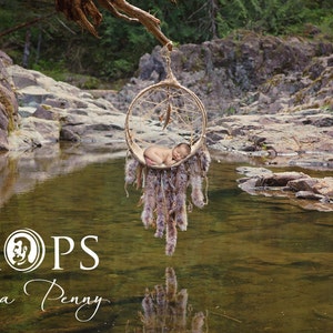 May include: A newborn baby rests in a hanging dream catcher prop, suspended over a tranquil stream. The dream catcher is adorned with feathers and fur, creating a natural, rustic aesthetic. The image includes the text "PROPS On a Penny".