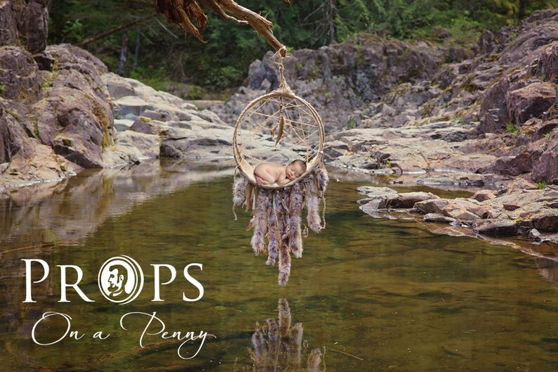 May include: A newborn baby rests in a hanging dream catcher prop, suspended over a tranquil stream. The dream catcher is adorned with feathers and fur, creating a natural, rustic aesthetic. The image includes the text "PROPS On a Penny".