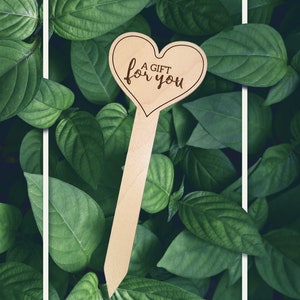 May include: A wooden heart-shaped plant marker with the text "A Gift for You" engraved on it. The marker is surrounded by green leaves.