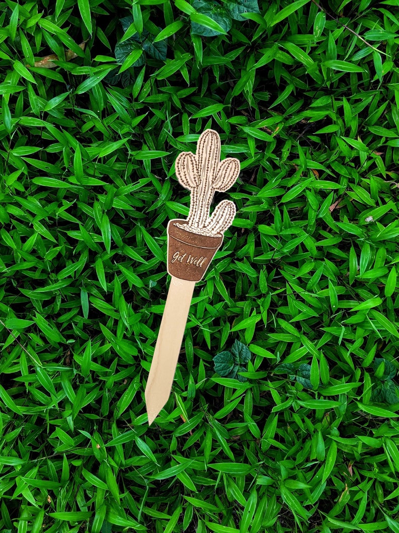 May include: Wooden get well soon card with a cactus design. The card is shaped like a stick and has the words "Get Well" written on it.