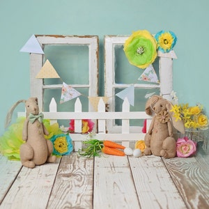 May include: Two brown bunny plushes with white accents sit in front of a white picket fence with a blue and green floral backdrop. The fence is decorated with a string of pastel blue and white triangle flags. Two carrots and two Easter eggs are in the foreground.
