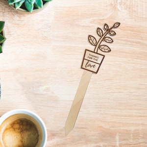 May include: Wooden plant marker with a brown stem and green leaves. The marker has a rectangular sign that reads "Thanks for the love".