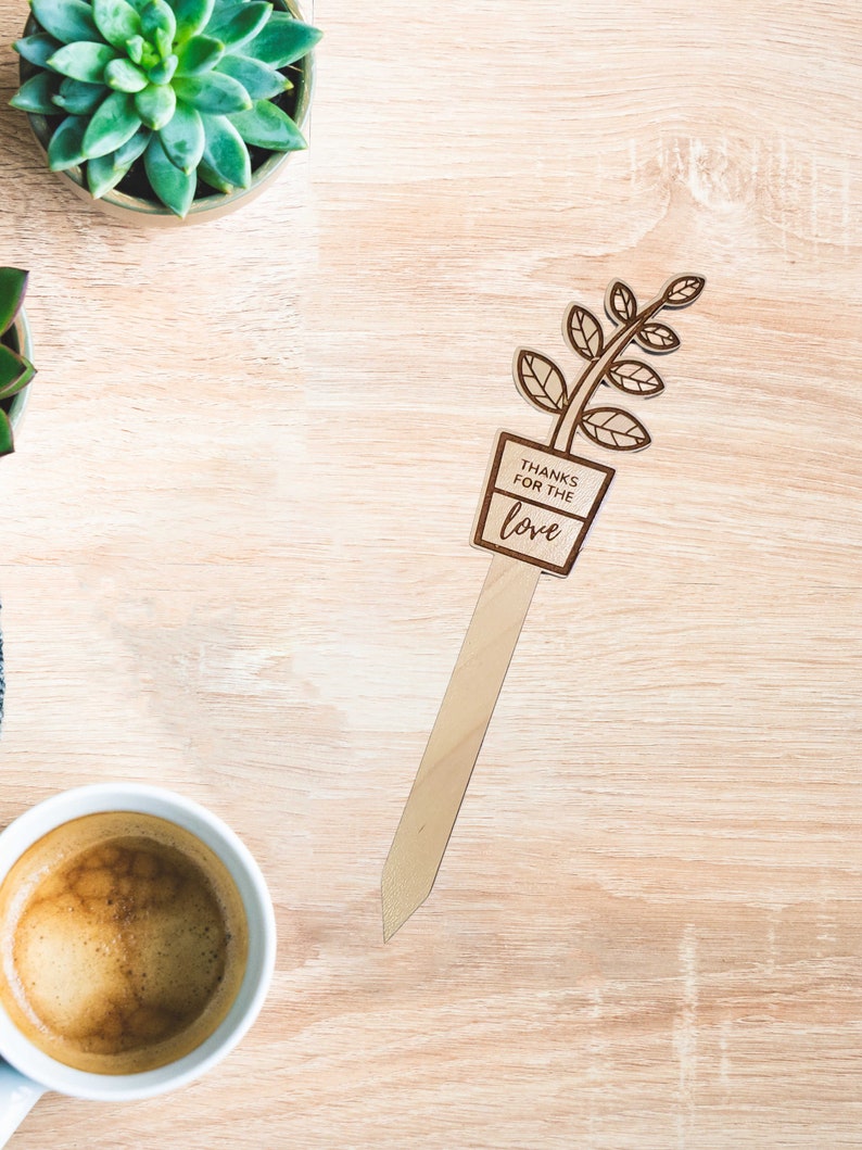 May include: Wooden plant marker with a brown stem and green leaves. The marker has a rectangular sign that reads "Thanks for the love".